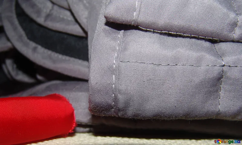 Detail šedé látky se švem a červeným okrajem v popředí.